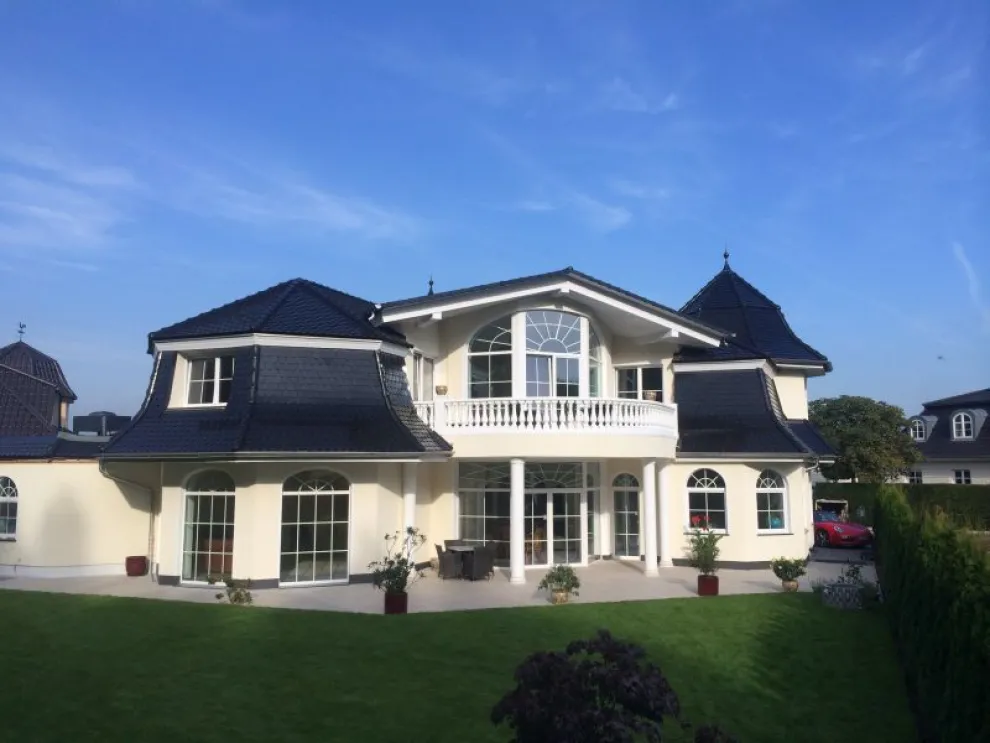 Die hellgelbe Hausfassade zusammen mit dem einladenden Balkon sowie dem vulkanschwarzen Biberschwanzziegel Rundschnitt lässt eine imposante Villa entstehen. Referenz: Fa. Longinus Jaeger-Bedachungen, Unna